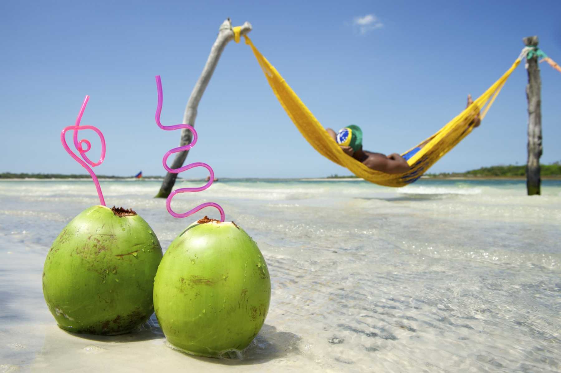 Two green coconuts with straws on the sand, and a person resting in a yellow hammock strung over shallow water.