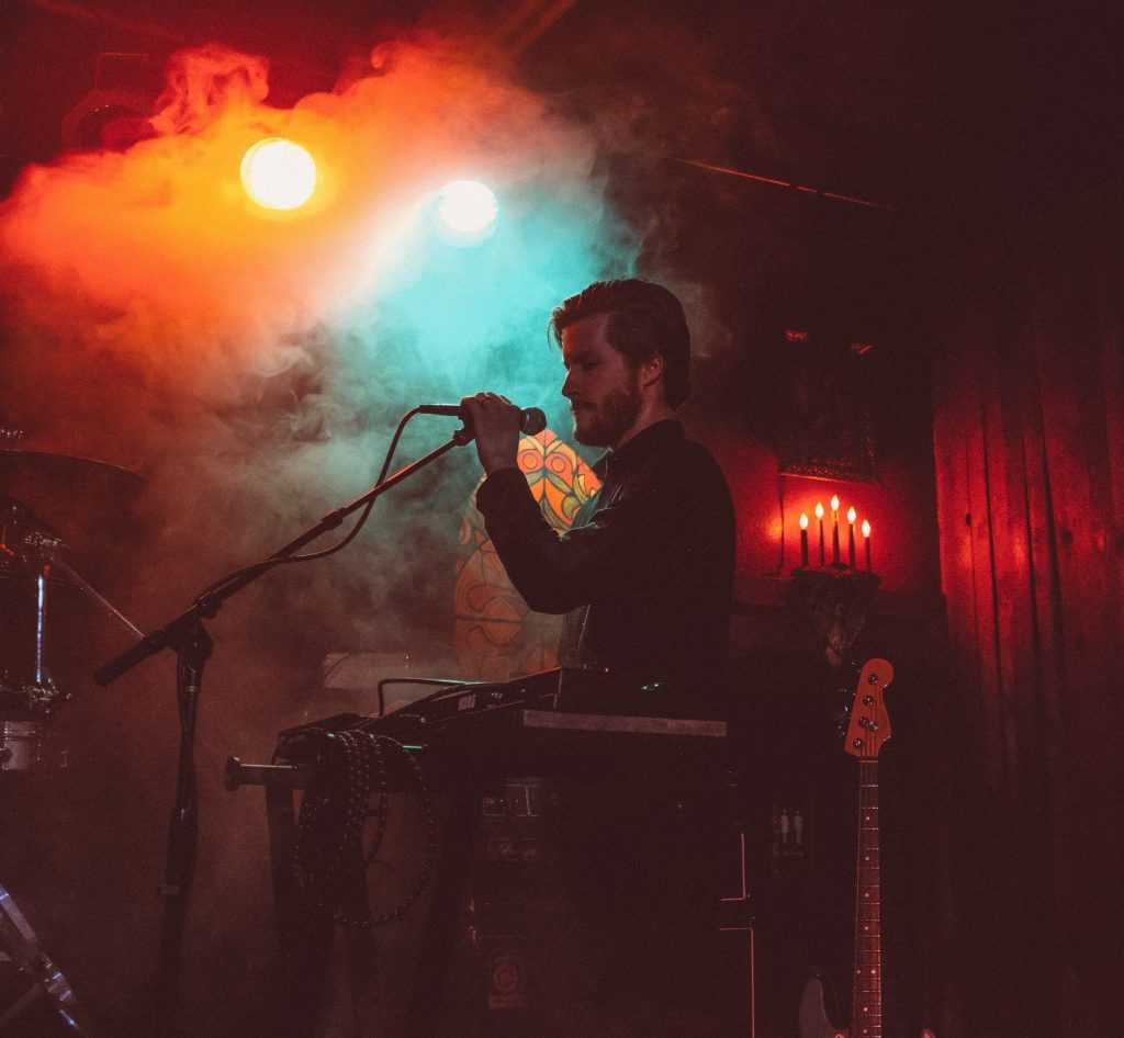 A singer at the microphone behind a keyboard, lit by red and teal stage lights in a smoky club.