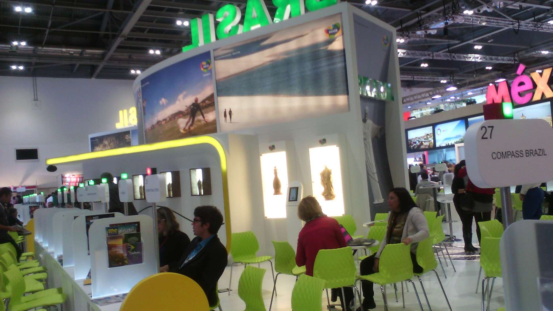 An exhibition stand with large screens and green seating on a busy trade show floor.