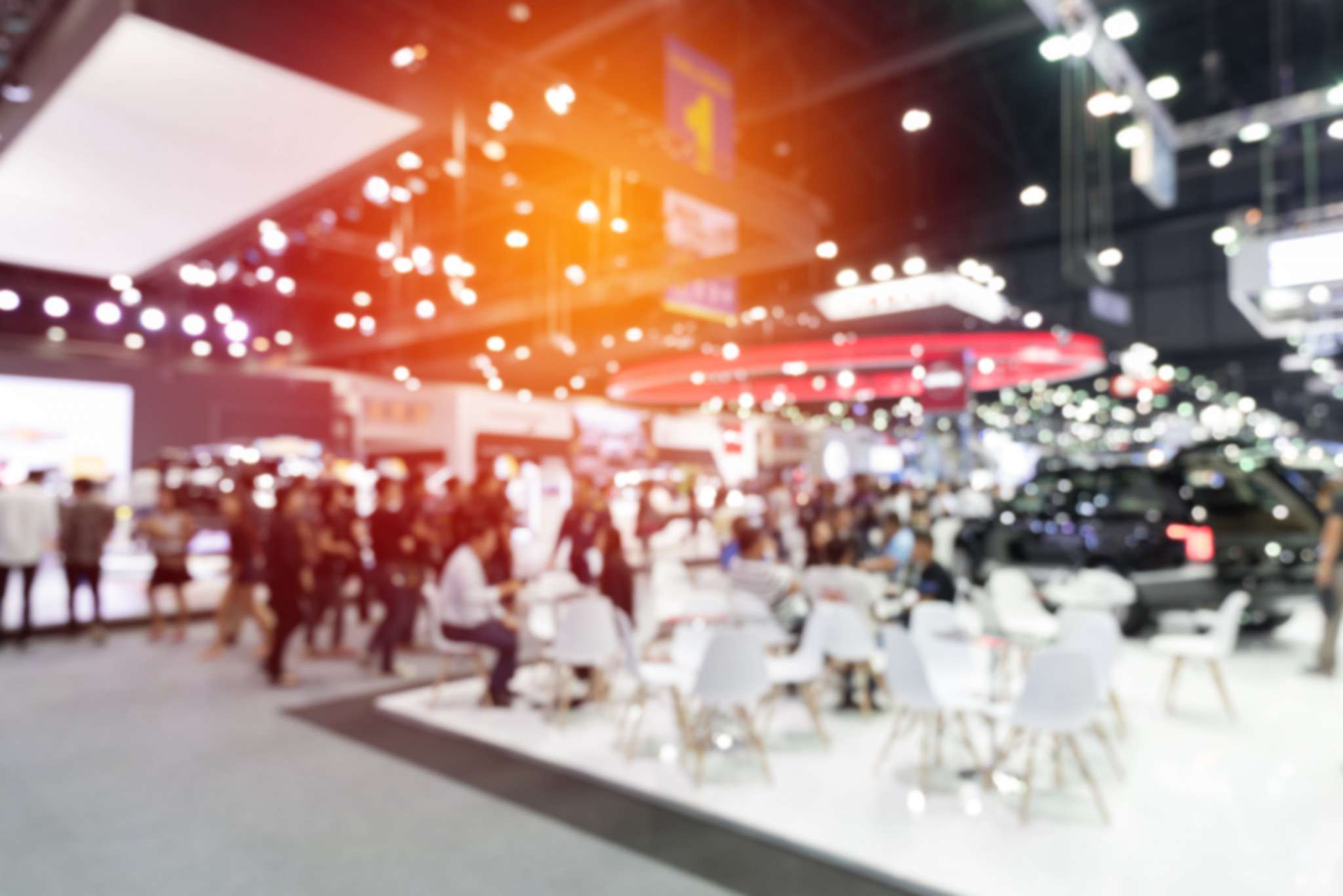Crowds moving between lit stands at a busy trade fair, with white tables and chairs in the foreground.