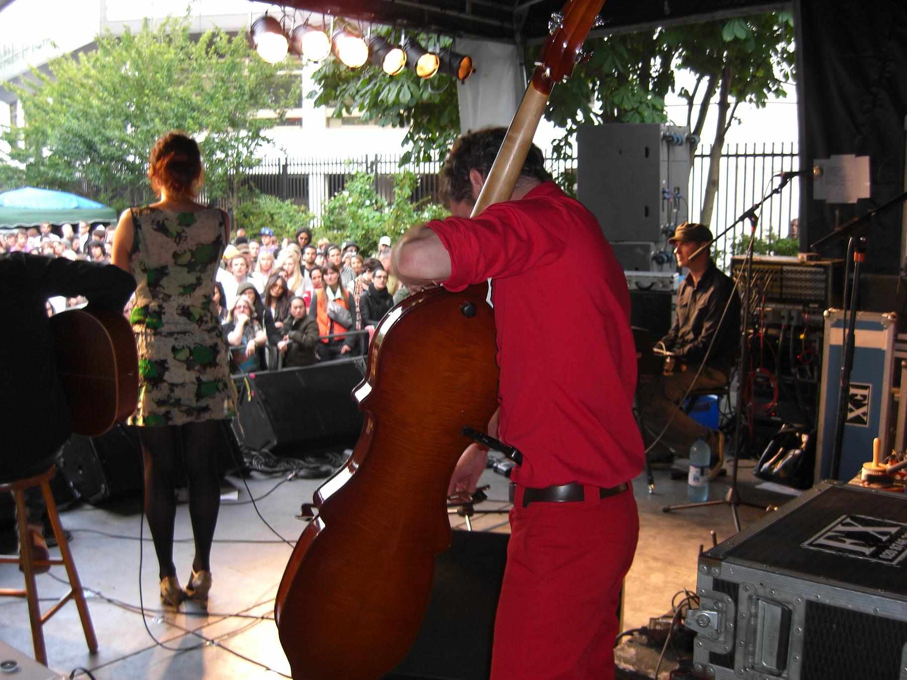 A double bass player and his band performing outdoors to a standing crowd at a festival.