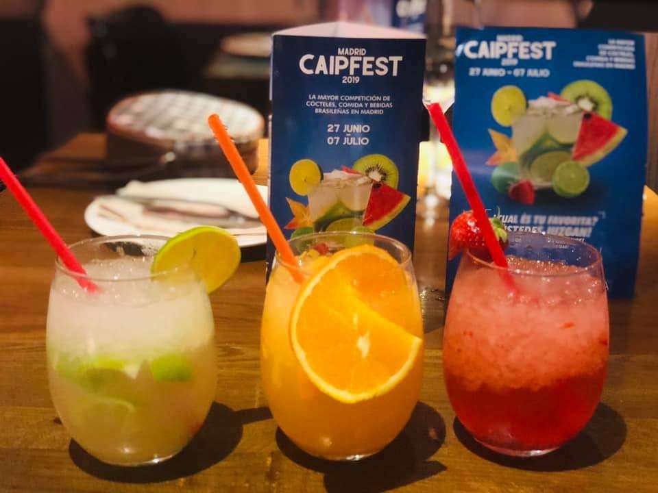 Three cocktails on a counter in front of posters for the Caipfest festival in Madrid.