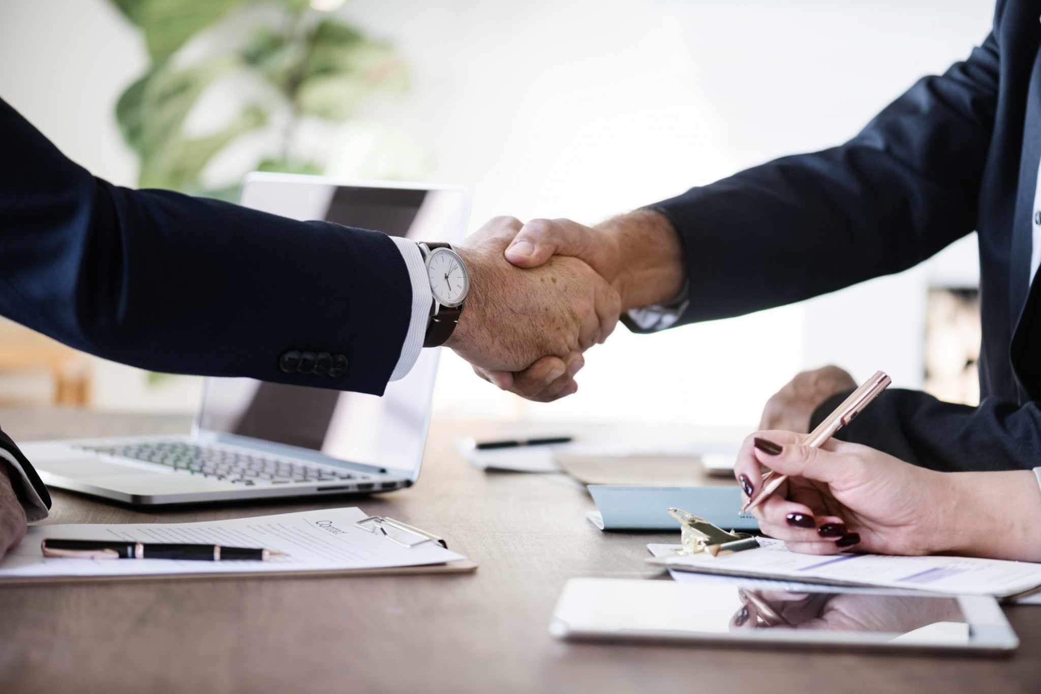 Two people in suits shaking hands across a desk at the close of a meeting.