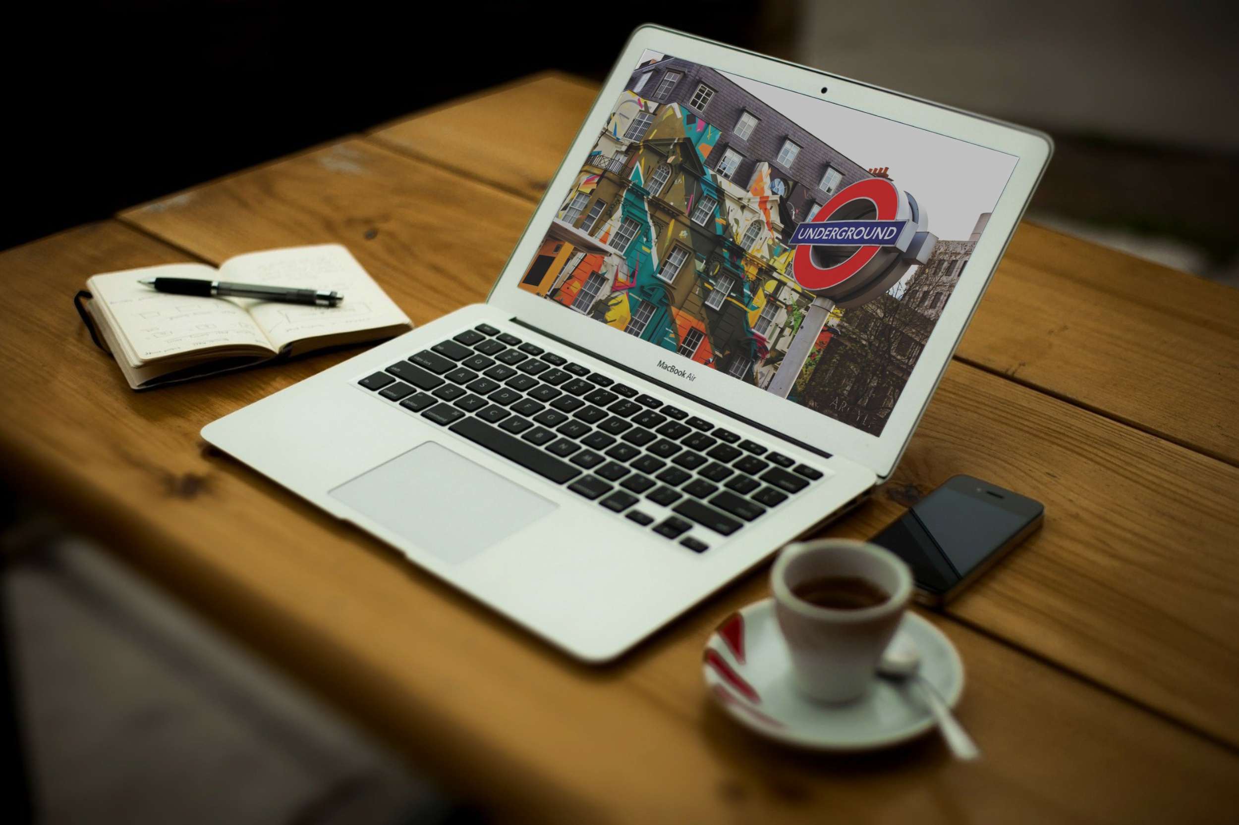 A laptop on a wooden table showing a London travel website, beside a notebook and an espresso.