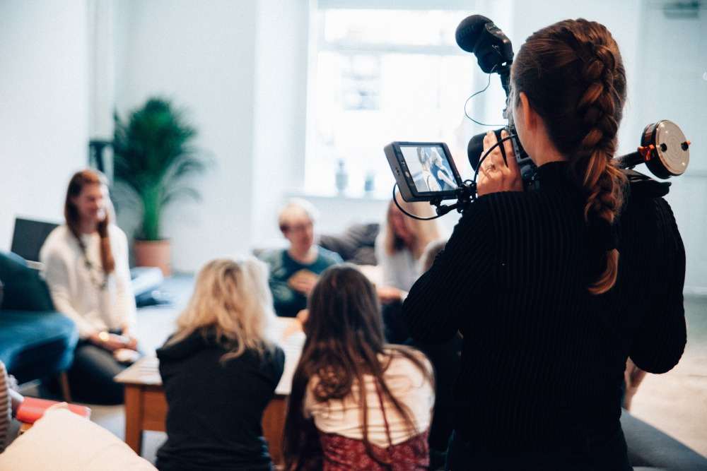 A camera operator filming a group being interviewed in a bright, informal room.