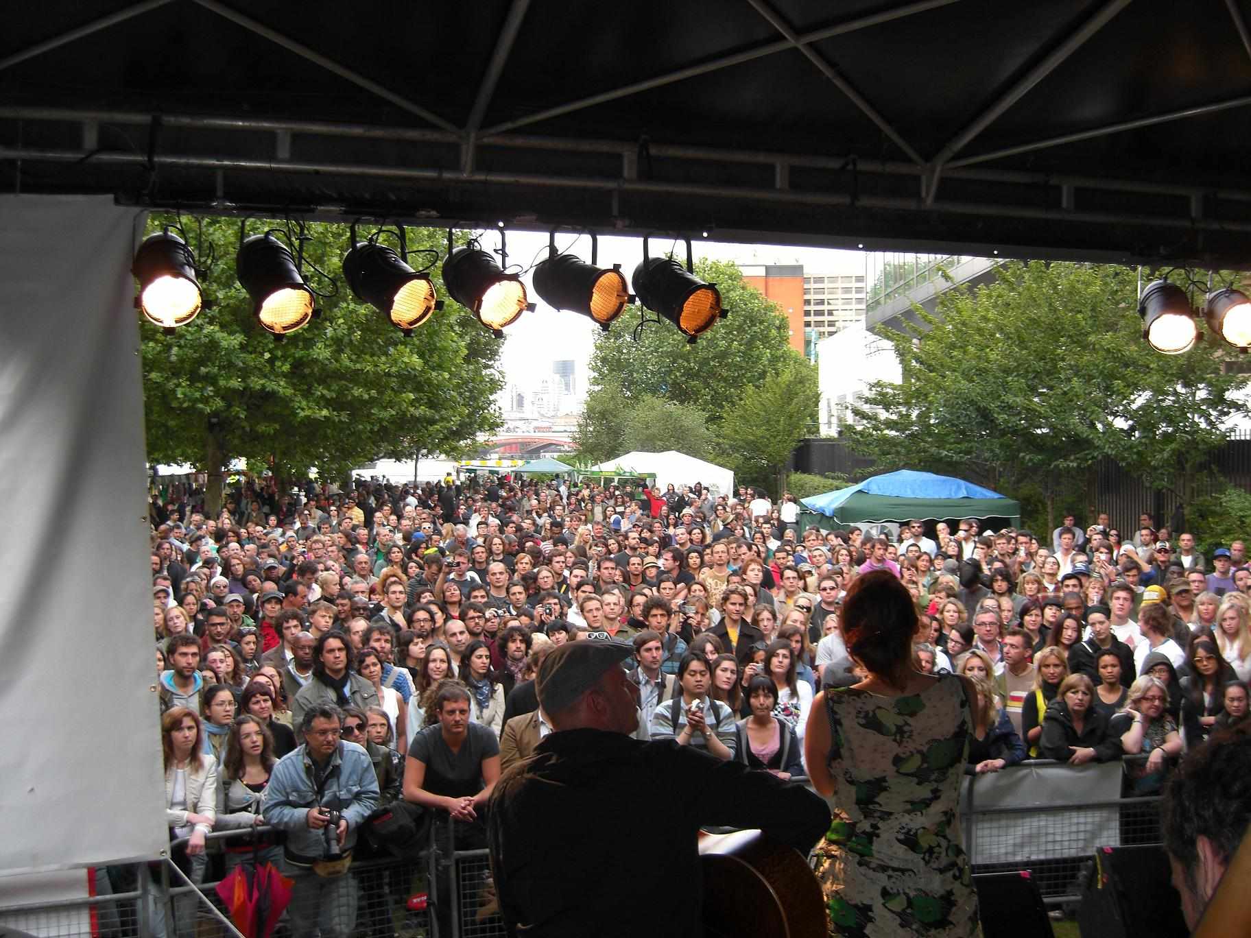 A large crowd watching a band play under strings of festoon lights at an open-air festival.
