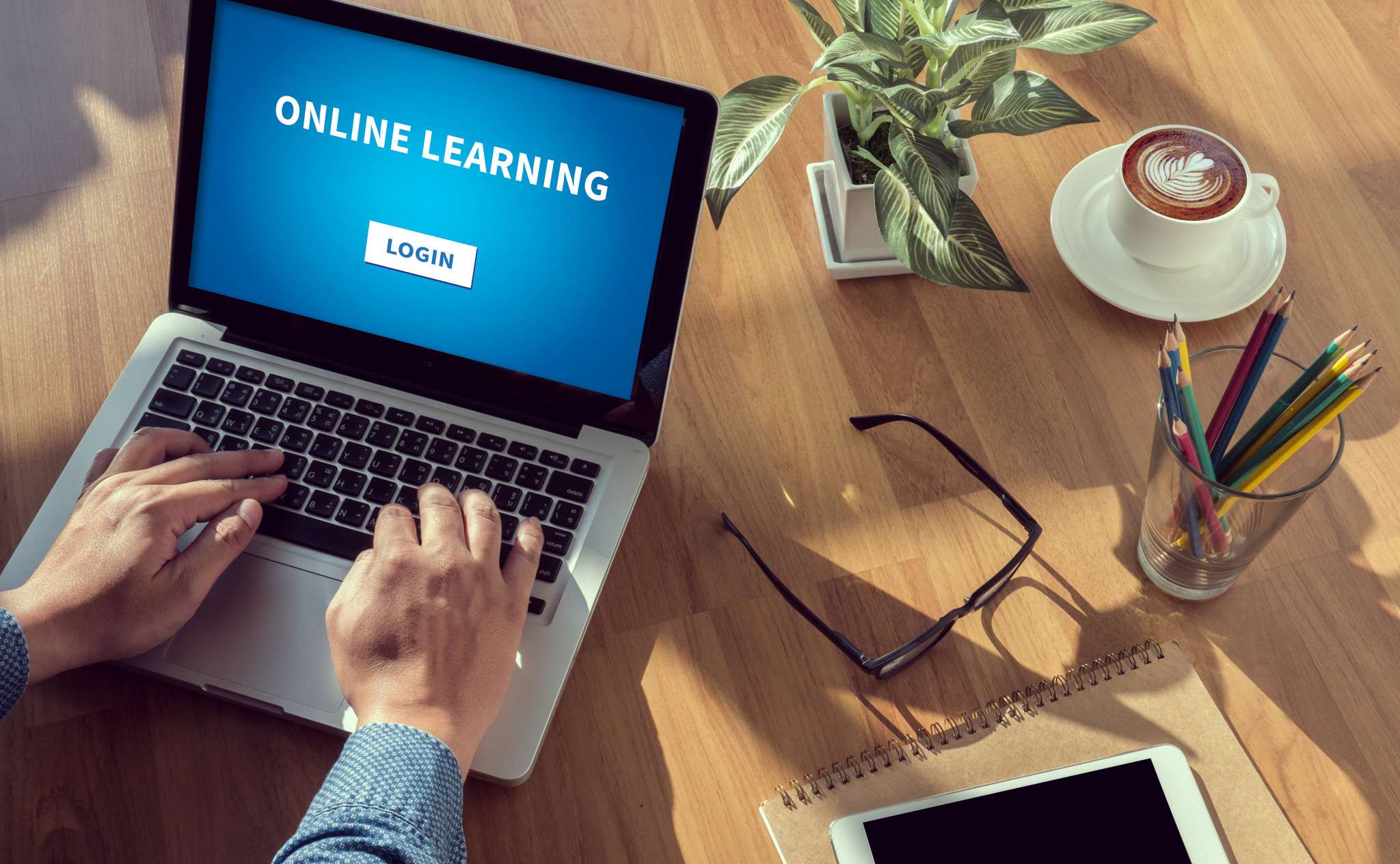 Hands typing on a laptop showing an online learning login screen, next to a notebook and a coffee.