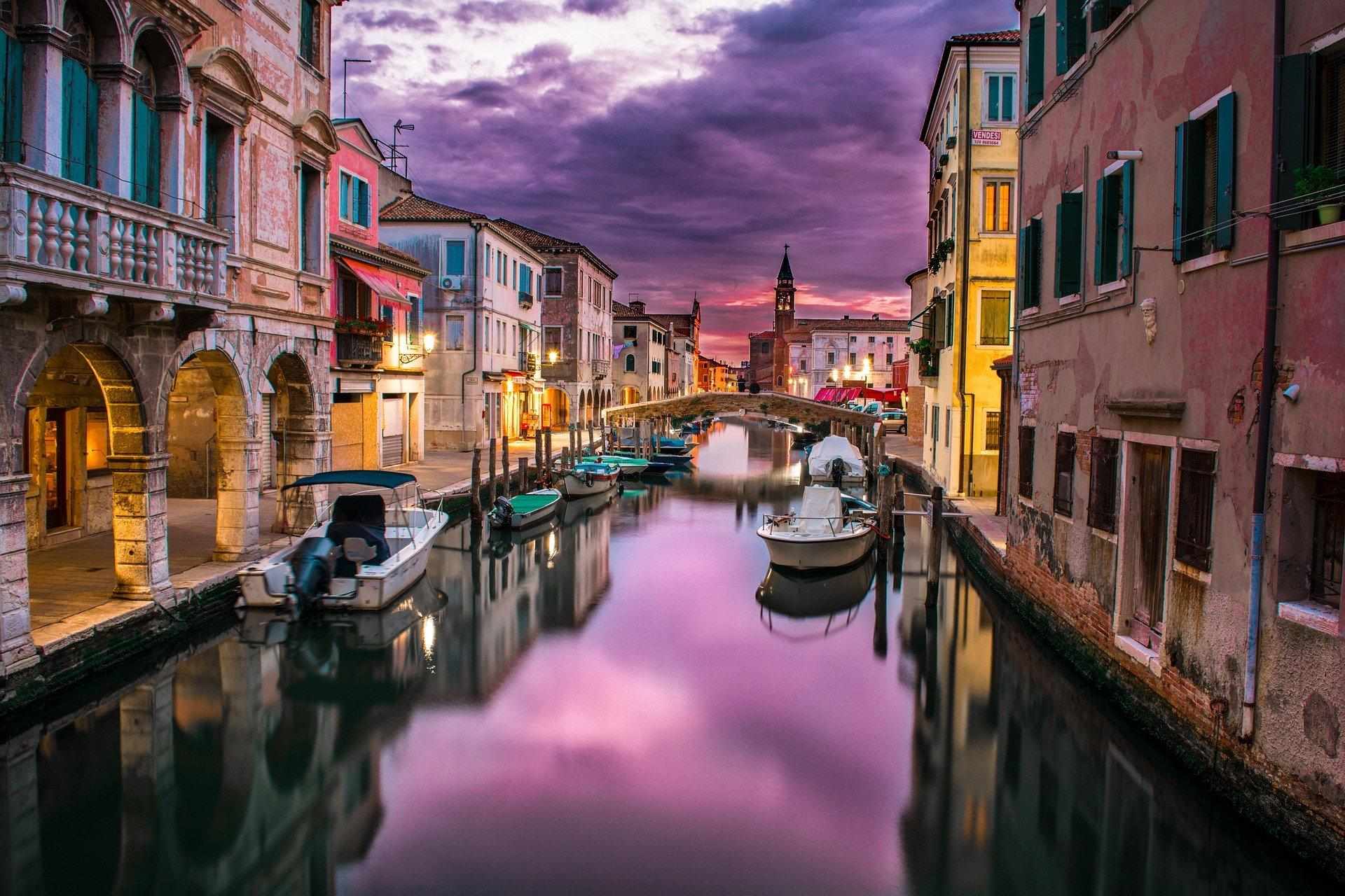 A canal at dusk between rows of old houses, with small boats moored along both banks.