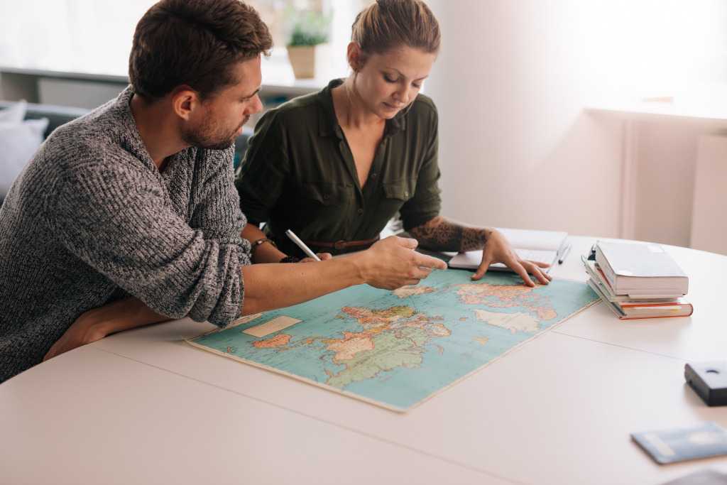 Two people leaning over a world map spread across a white table, one of them pointing at Europe.